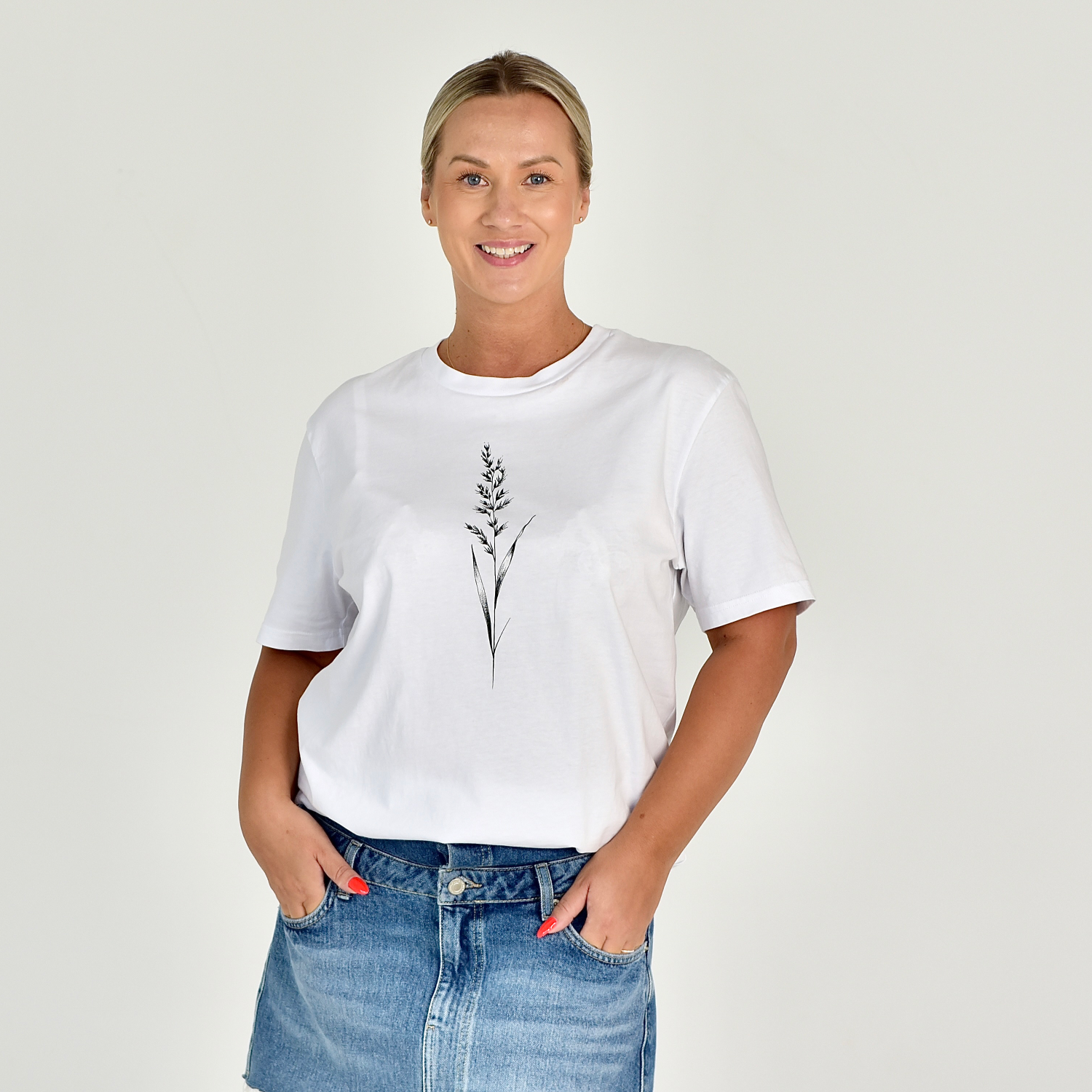 Stilingi balti moteriški marškinėliai su minimalistiniu smilgos piešiniu, demonstruojami ant besišypsančios moters. Puikus pasirinkimas kasdienai ir laisvalaikiui. Designedbyme.lt originalus dizainas, Moteriški marškinėliai, Vasaros kolekcija, Populiariausi moteriški marškinėliai 2024, BESTSELLER'iai, Naujausi dizainai