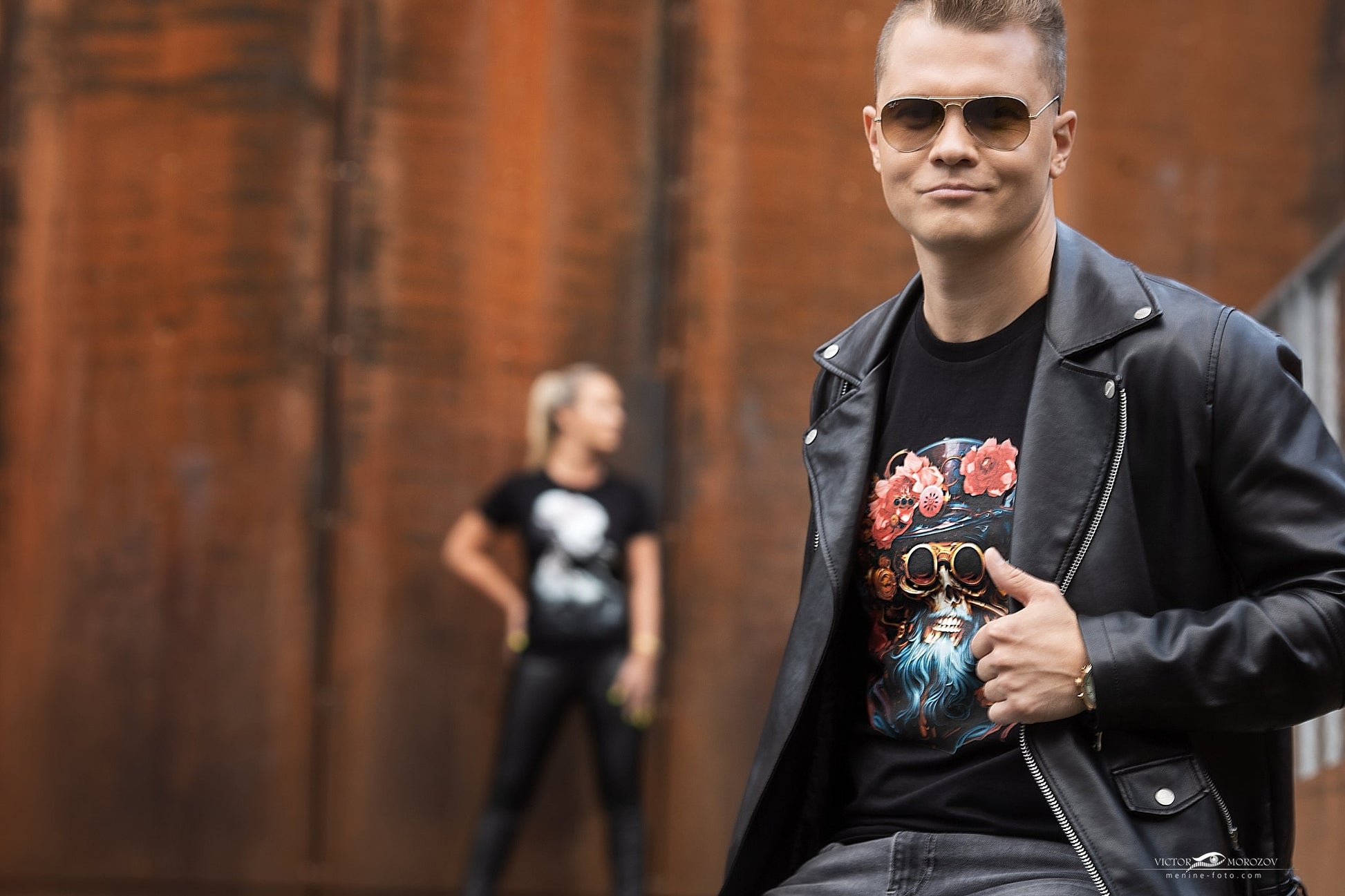 Vyriškis su juoda odine striuke vilki "Captain Jack" marškinėlius su piratišku dizainu, kuriuose dominuoja gėles ir akinius vaizduojantis spalvingas piešinys. Fone matosi moteris su panašia apranga, Designedbyme.lt, Designed By Me, marškinėliai, užrašai ant marškinėlių, maikutes vyrams, maikutės vyrams, vyriskos maikes, vyriskos maikutės, Vyriški marškinėliai,Visi produktai