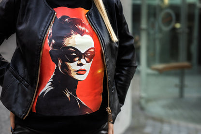 Stilingi moteriški marškinėliai su Catwoman atvaizdu, ryškiomis spalvomis ant raudono fono. Marškinėliai dėvimi po juoda odinė striuke, atspindintys modernų ir drąsų stilių. Puikus pasirinkimas Designedbyme.lt gerbėjoms, Moteriški marškinėliai, Naujausi dizainai