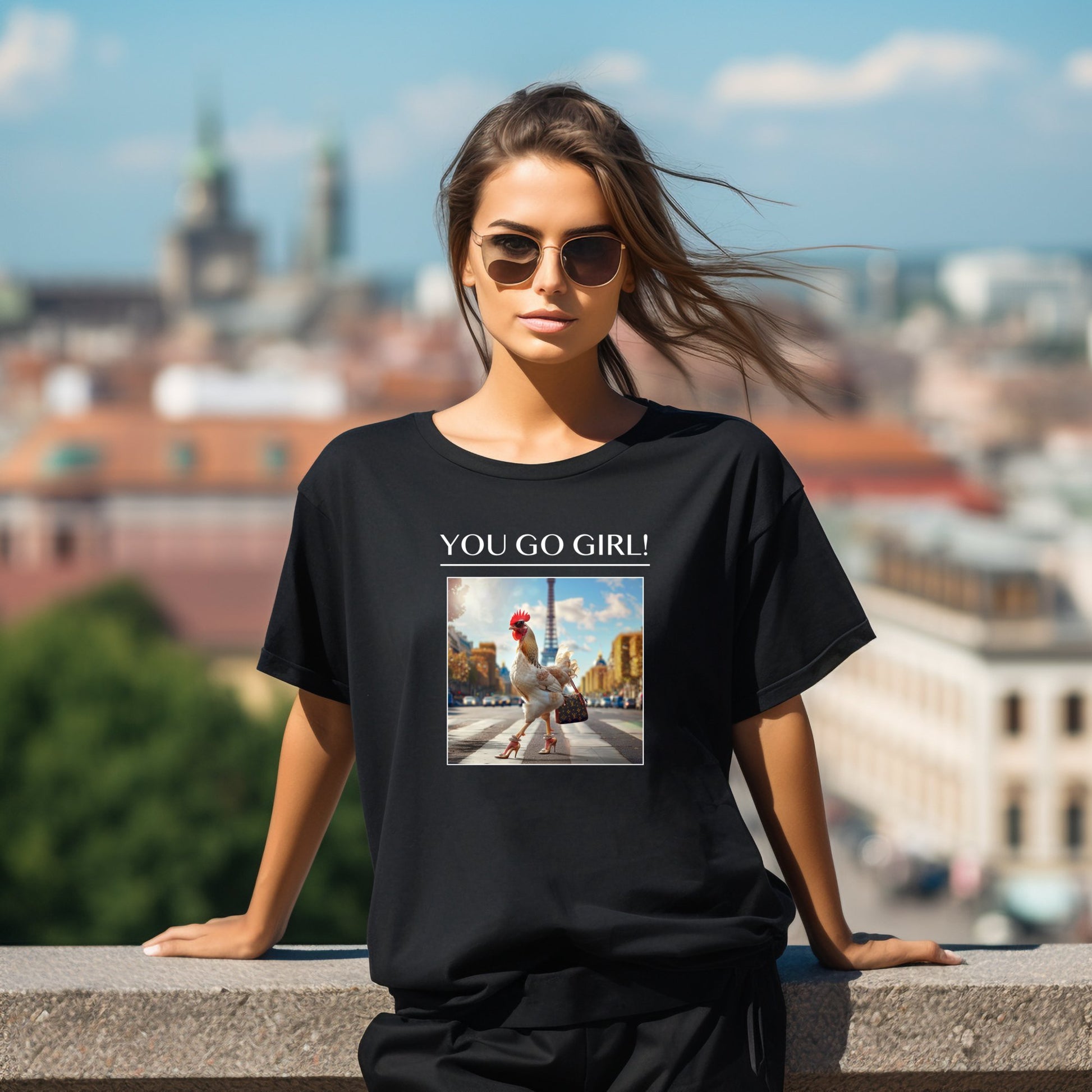 Stilingi juodi moteriški marškinėliai su originaliu "You Go, Girl!" užrašu ir vištytės paveikslėliu miesto fone, dėvimi jaunos moters su akiniais nuo saulės miesto panoramos fone. Unikalus Designedbyme.lt dizainas, Moteriški marškinėliai, Vasaros kolekcija , Naujausi dizainai